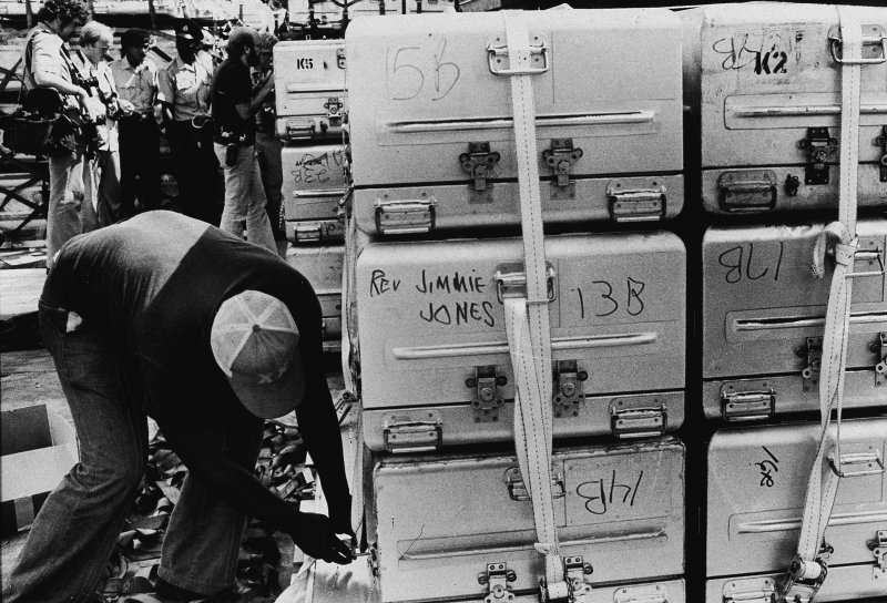 An unidentified man a strap onto a stack of aluminum coffins for shipment to the United States, following the more than 900 deaths in the mass suicide staged in Jonestown by members of the People's Temple and their leader, the Reverend Jim Jones, Georgetown, Guyana, Nov. 23, 1978. A group of photographers and police officers stand in the background.